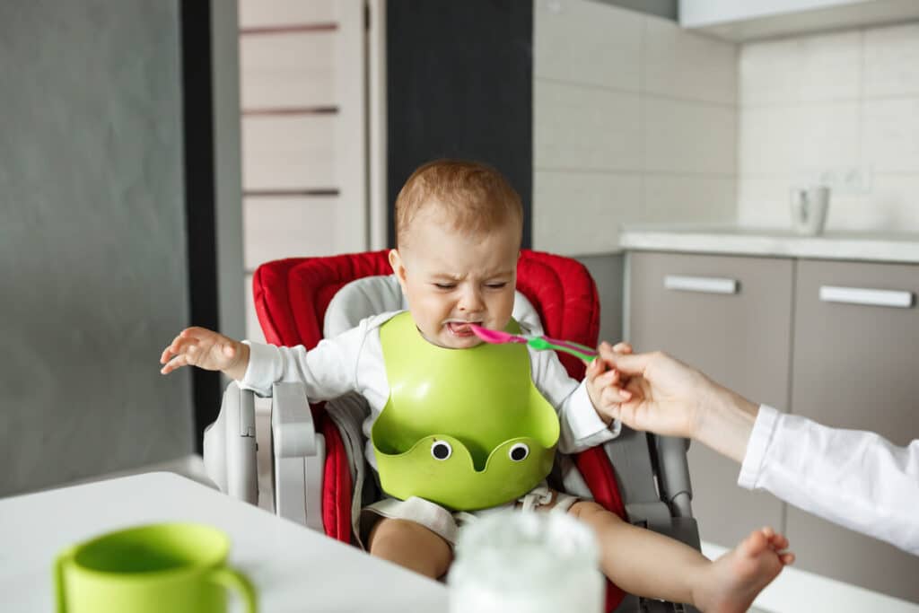 Bébé pleure en mangeant sa purée : causes, conseils et solutions 2 Bébé assis dans sa chaise haute refusant la cuillère de purée en pleurant.