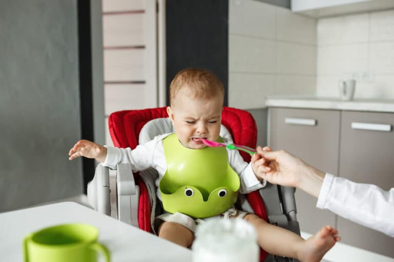 Bébé assis dans sa chaise haute refusant la cuillère de purée en pleurant.
