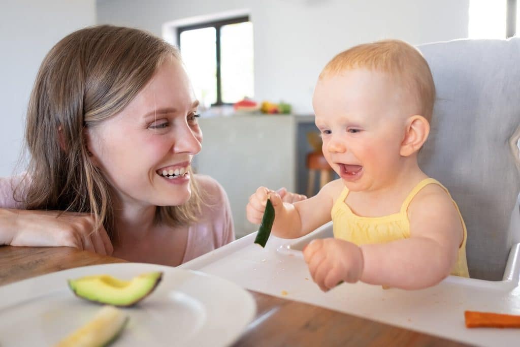 Bébé pleure en mangeant sa purée : causes, conseils et solutions 3 Bébé souriant touchant des légumes cuits avec ses mains lors d'un repas.