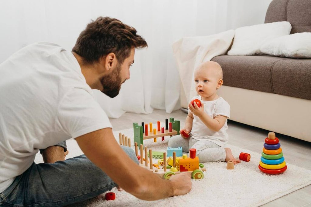 Parent encourageant son bébé allongé sur le ventre sur un tapis de sol à attraper un jouet.
